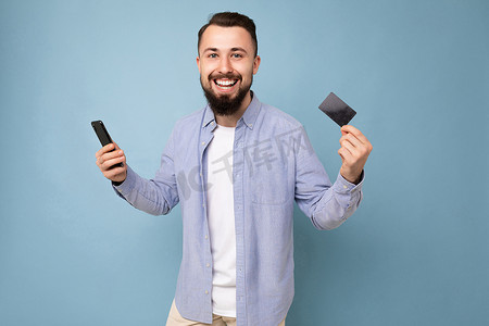 照片中，穿着休闲蓝色衬衫和白色 T 恤的帅气迷人、微笑的黑发未剃须年轻人穿着蓝色背景墙，拿着信用卡和手机
