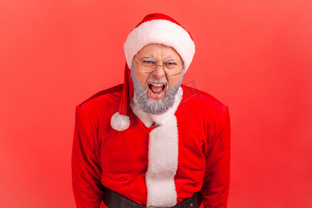 愤怒的老人留着灰色的胡须，穿着圣诞老人的服装，用咄咄逼人的表情大声尖叫，看着镜头，大喊大叫。