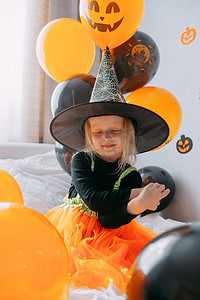 儿童万圣节 — 一个女孩戴着女巫帽，穿着嘉年华服装，家里有通风的橙色和黑色气球。