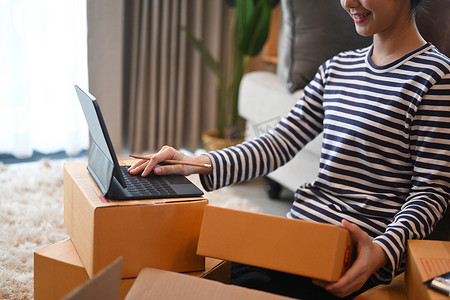 微笑的年轻女子在电脑平板电脑上确认顾客的订单，并准备包裹箱以供送货。