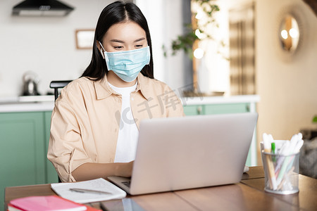 戴着医用口罩的亚洲女孩在感染新冠病毒时在家工作，使用笔记本电脑
