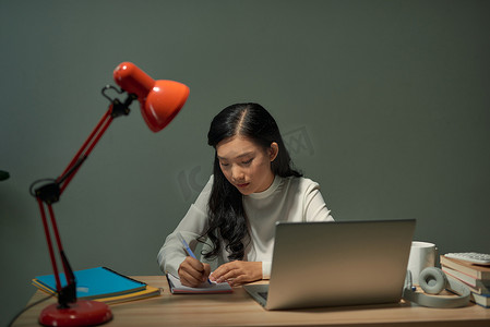 漂亮的年轻学生坐在办公桌前做作业，她正在用笔记本电脑连接到互联网