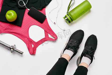 健身俱乐部里，穿着黑色运动鞋的女孩腿坐在垫子上，周围放着手机、耳机、毛巾和摇床。