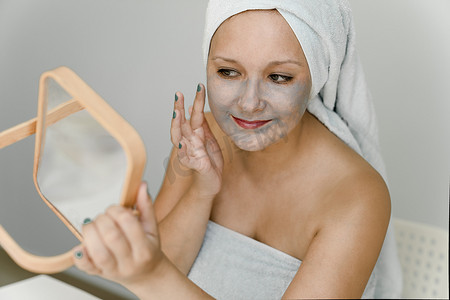 年轻女子一边照镜子，一边把灰色的化妆品粘土涂在脸上，头发和身体都裹着毛巾。