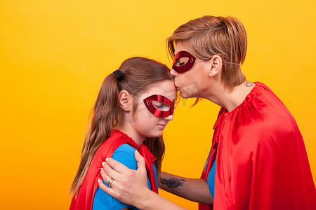 孩子超人摄影照片_穿着超人服装的母亲在黄色背景中亲吻他的小女孩额头