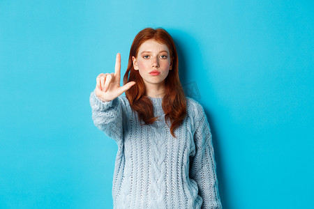 穿毛衣的严肃红发女孩摆出禁忌姿势，伸出一根手指，摇晃食指表示不赞成、反对和禁止某事，蓝色背景