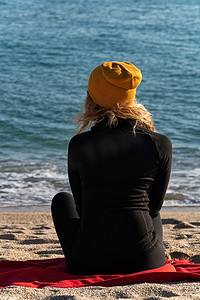 一个穿着黑色运动夹克、戴着黄色帽子、金发卷发的女孩坐在沙滩上，看着蓝色的大海。