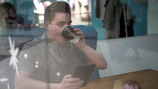 一个年轻人坐在咖啡馆的玻璃杯后面，手里拿着一台平板电脑。