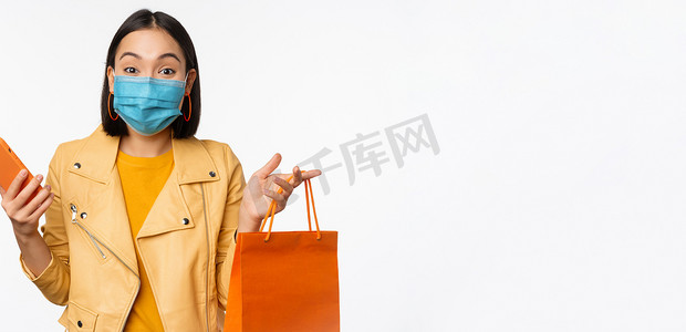 时尚亚洲女购物者的形象，手持智能手机和没有商店标志的购物袋，戴着 covid-19 的医用面罩，白色背景
