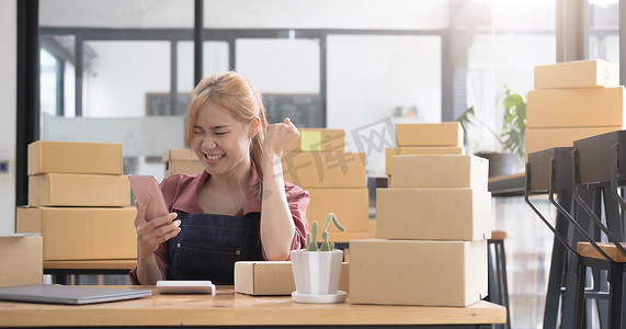 快乐的年轻亚洲女企业家，在家庭办公室用智能手机查看在线购物商店的订单后，为销售成功而微笑，在线商业和电子商务的概念