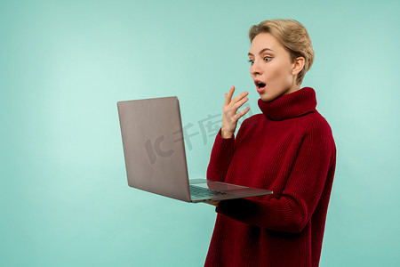 穿着毛衣的金发美女惊讶地看着笔记本电脑屏幕，蓝色背景上张着嘴