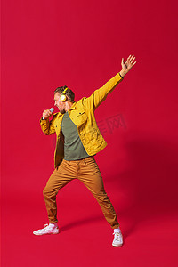 红色乐谱摄影照片_唱歌的年轻人用一只手享受歌曲跳舞，使用电话和无线耳机，穿着牛仔裤黄色夹克，隔离在红色背景中。