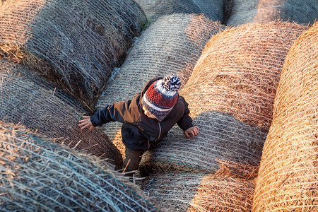 一个戴着针织帽的可爱男孩爬到一堆稻草堆的顶部。