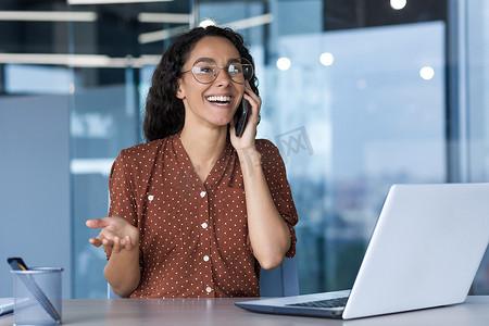 年轻成功的西班牙裔女商人在现代办公室工作，女员工愉快地通过电话与同事交谈并微笑，坐在办公桌前使用笔记本电脑