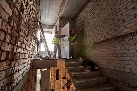 两名身穿工作服、头戴安全帽的工人正在一栋在建高层建筑未完工的楼梯间安装门