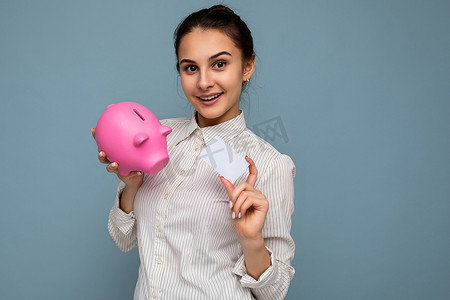 快乐积极微笑的年轻美丽漂亮黑发女人的肖像，带着真诚的情感，穿着休闲白衬衫，隔离在蓝色背景上，有复制空间，拿着粉红色的小猪盒，装钱和信用卡付款。