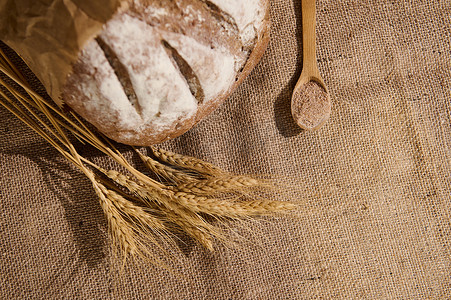 一条全麦面包，撒上面粉、小麦小穗和麻袋桌布上的木勺，上面有麸皮