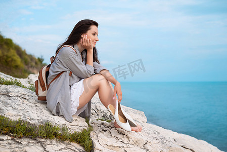 一个漂亮的旅游女孩，背着时尚的背包，穿着夏装，坐在海边的一块白色岩石上
