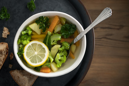 木桌上金属托盘上的一个轻碗里，放着胡萝卜、西兰花和欧芹的素食蔬菜汤