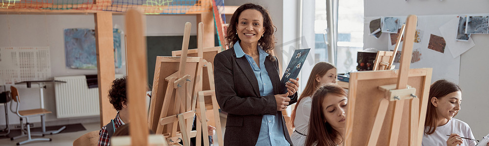 现代课堂上微笑的美国女教师与孩子们在背景中画课的画像