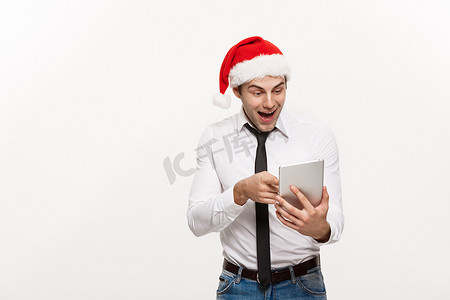 圣诞节概念-英俊的商人戴圣诞老人的帽子玩平板电脑与令人惊讶的面部表情。
