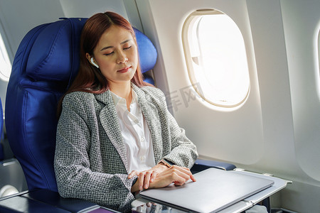 在飞机上睡觉摄影照片_坐在商务舱的飞机上穿着正式西装的成功亚洲女商人或企业家的肖像。