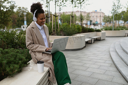 非洲裔美国年轻女性戴着耳机在笔记本电脑上在线工作，在城市背景下喝着一杯咖啡，脸上挂着微笑，这是听音乐播客的概念