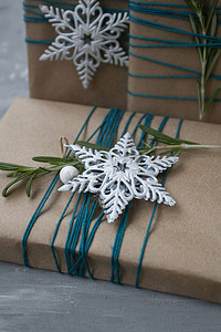 灰色枝条摄影照片_三件用牛皮纸包裹的圣诞礼物，用蓝色线包裹，灰色混凝土背景上有白色雪花和迷迭香枝条。
