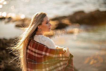 迷人的金发白种女人在日落时分在海滩上享受时光，坐在毯子里，看着一边，背景是落日的天空和大海。