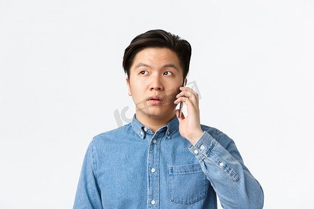 优柔寡断的亚洲男人在打电话时发呆、交谈、困惑地看着别处、站在白色背景上的特写镜头。