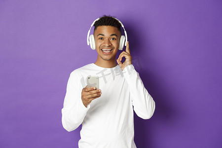 英俊的非洲裔美国男子微笑着，戴着耳机听音乐，拿着智能手机，站在紫色背景上的肖像