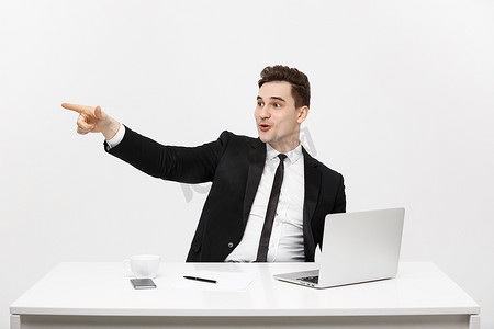 经营理念：穿着西装的英俊商人坐在办公室里，手指指向复印空间和灰色背景中隔离的笔记本电脑的肖像