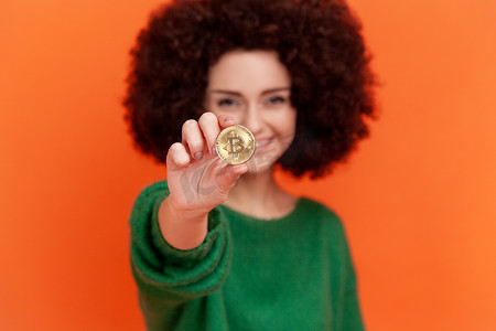 身穿绿色休闲毛衣、留着非洲发型、心满意足的女性拿着黄金比特币，面露露齿微笑地看着相机，这是区块链概念。