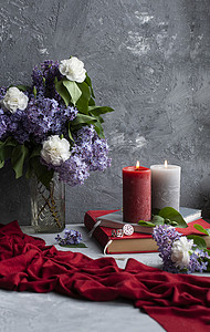 红色大气背景摄影照片_大气照片：灰色背景中的一束丁香和康乃馨、几支大蜡烛、书籍和珠宝