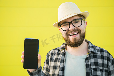 快乐微笑、留着胡子、戴着帽子的男人在黄色背景上展示手机 — 空荡荡的空间和广告场所