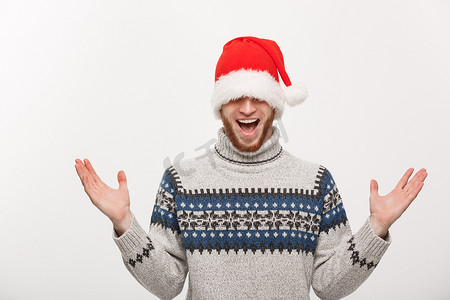 圣诞老人胡子帽子摄影照片_假日概念-穿着毛衣的年轻胡子男人喜欢玩圣诞老人的帽子。