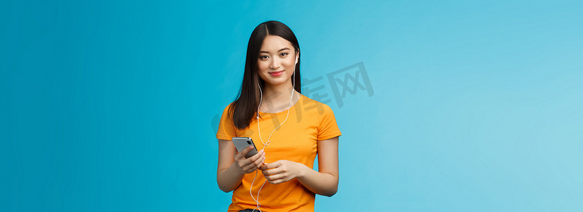 无忧无虑、迷人、温柔的亚洲城市女性戴着耳机，通过耳机听音乐，拿着智能手机，微笑着高兴，发现了很棒的新歌，站在蓝色背景