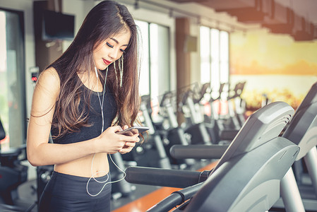 行走的力量摄影照片_亚洲女性在跑步机上的健身房锻炼或力量训练时使用智能手机。