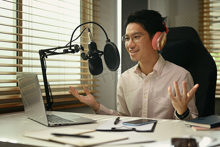 播摄影照片_积极的亚洲男子电台主持人使用电容麦克风和笔记本电脑在小型家庭工作室录制播客