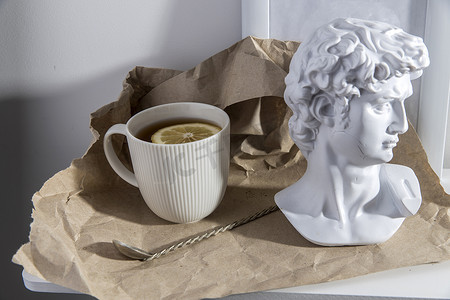 棕色工艺纸上有一个带茶和柠檬的白色瓦楞纸杯，上面有阿波罗石膏头像和长柄上的白铜茶匙