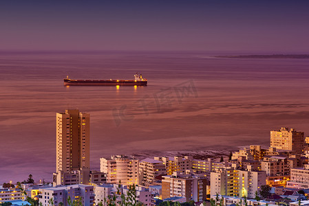 湾海城市大景摄影照片_夜间，在黑暗的天空下，一艘货船在平静的海洋上俯瞰摩天大楼和沿海城市。