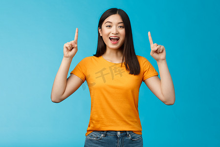 有趣的亚洲女孩向你介绍新产品分享促销，举起双手指指点点地微笑，看起来兴奋和乐观，热情的广告，站在蓝色背景