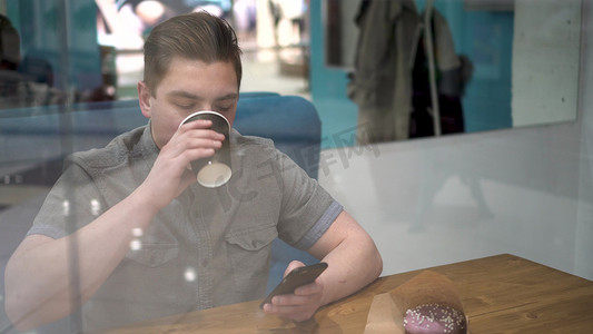 一名年轻人坐在咖啡馆的玻璃后面，手里拿着手机。
