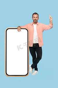 手势注意力指向中年灰发男子，倚在巨大的巨型智能手机上，白色屏幕在镜头前快乐地微笑着，穿着蓝色背景中突显的桃色衬衫