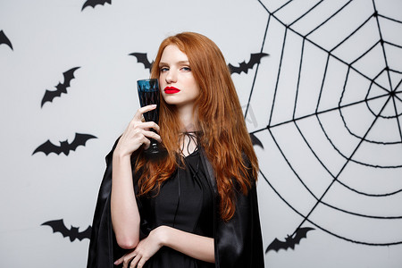 血淋淋的摄影照片_万圣节女巫概念 — 快乐的万圣节女巫在深灰色工作室背景上拿着一杯血淋淋的红酒，上面有蝙蝠和蜘蛛网。