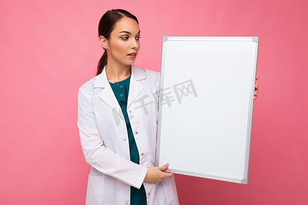 穿着白色医用外套的女医生拿着空白板，上面有粉红色背景上隔离的文本复制空间。