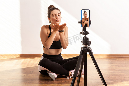 健身教练盘腿坐在三脚架上的手机摄像头前，送出飞吻。