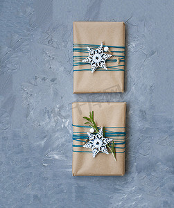 两份用牛皮纸包裹的圣诞礼物，用蓝色线包裹着，灰色混凝土背景上有白色雪花和迷迭香枝条。