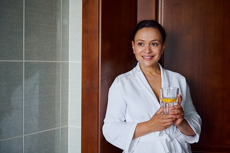 清爽浴室摄影照片_特写令人惊叹的多民族黑发女郎，皮肤干净健康，穿着浴袍，拿着柠檬水玻璃