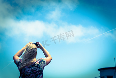 斜挎包摄影照片_背着斜挎包的金发白人女士用手机在蓝天上拍照